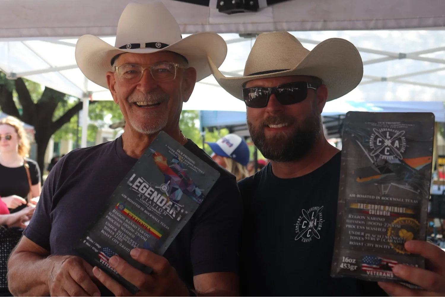 Two men in cowboy hats smiling under the Legendary Aviation Coffee tent, holding bags of Ethiopia and The Super Patriot Colombia single-origin coffee